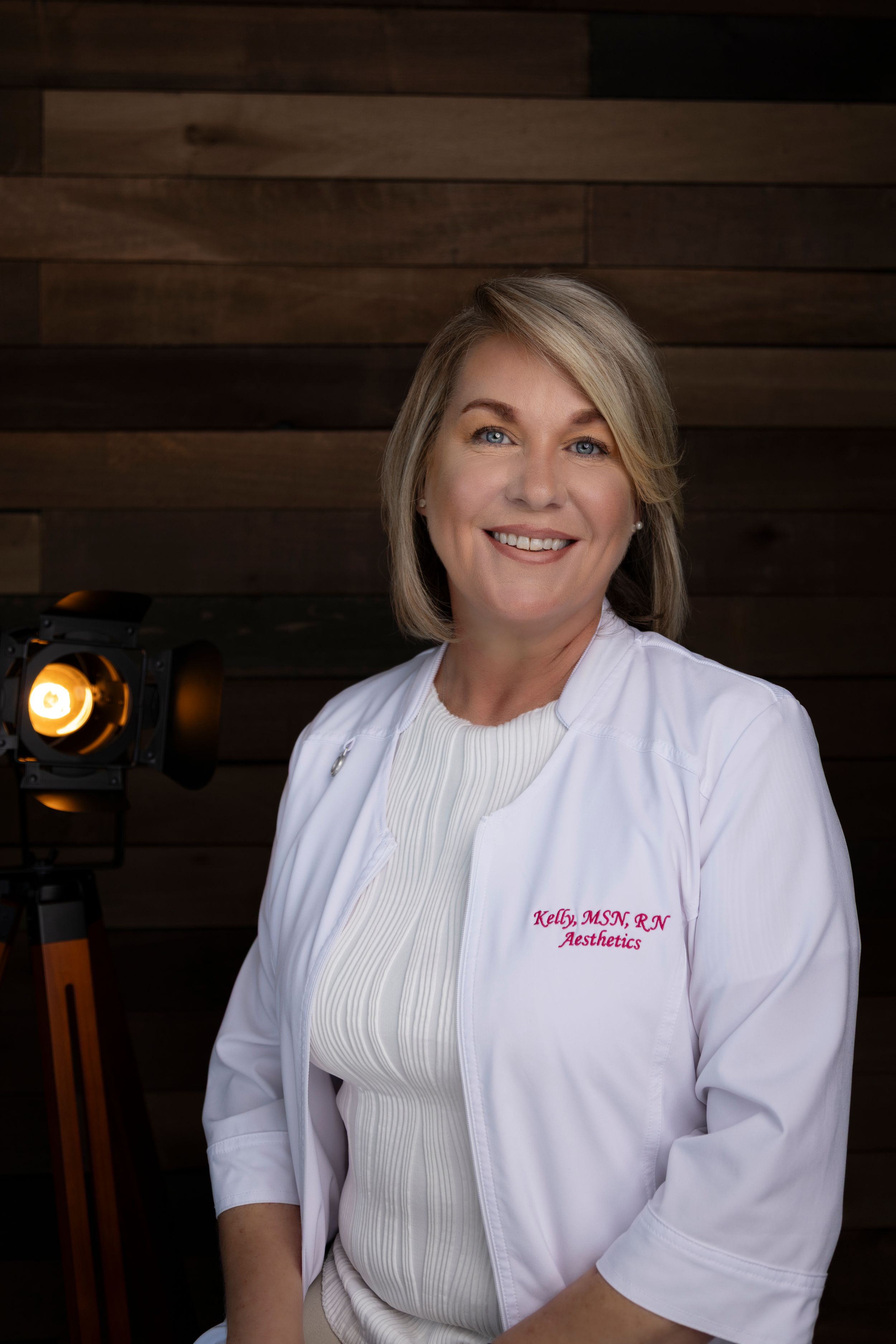 Healthcare professional in a white lab coat and ribbed sweater poses against wooden wall with studio lighting.