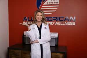 Medical professional in white coat standing against red wall with All American Weight Loss & Wellness signage.
