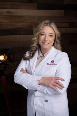 Healthcare professional wearing a white lab coat stands with crossed arms in front of dark wooden wall background.