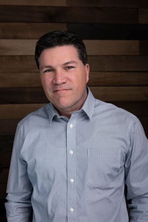 Professional headshot portrait of someone in a light blue button-down shirt against a wooden plank wall background.