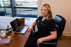 Healthcare professional in scrubs sits at desk with laptop in medical office setting overlooking waterfront.
