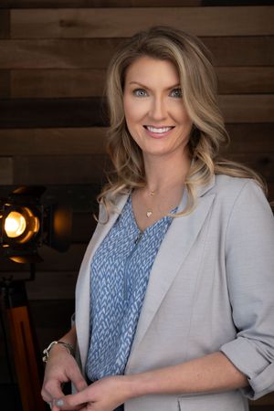 Professional headshot against rustic wood paneling showing a light gray blazer and patterned blue top.