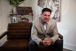 Professional wearing a beige suit sits in a rustic office with vintage decor and antique wall signage.