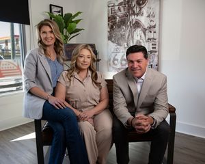 Three real estate professionals sit together on a modern couch in a bright office with Route 66 artwork on the wall.