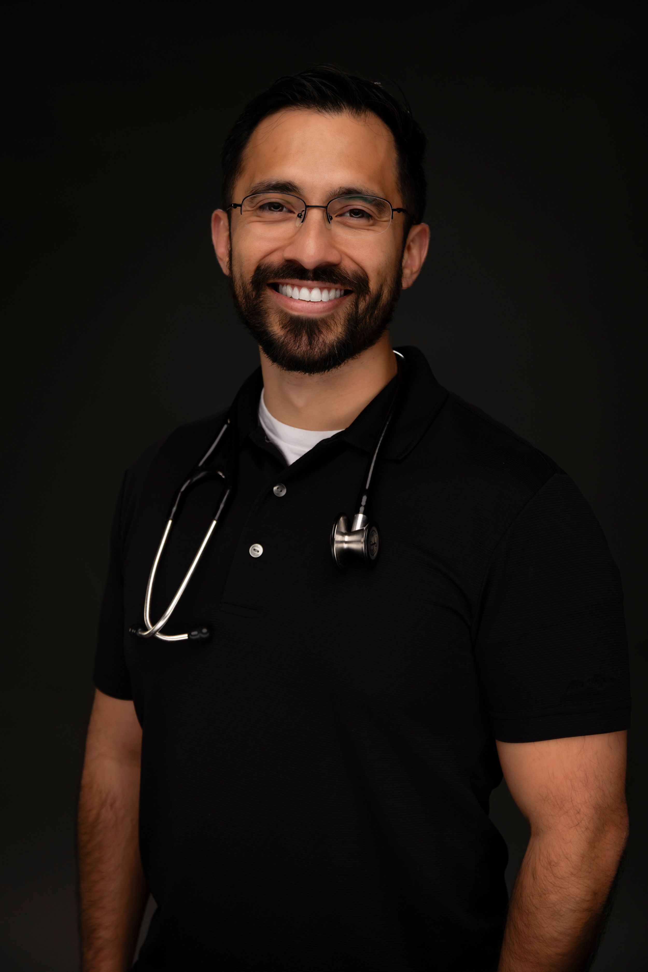 A healthcare professional in a black polo shirt wearing glasses and a stethoscope poses against a dark background.