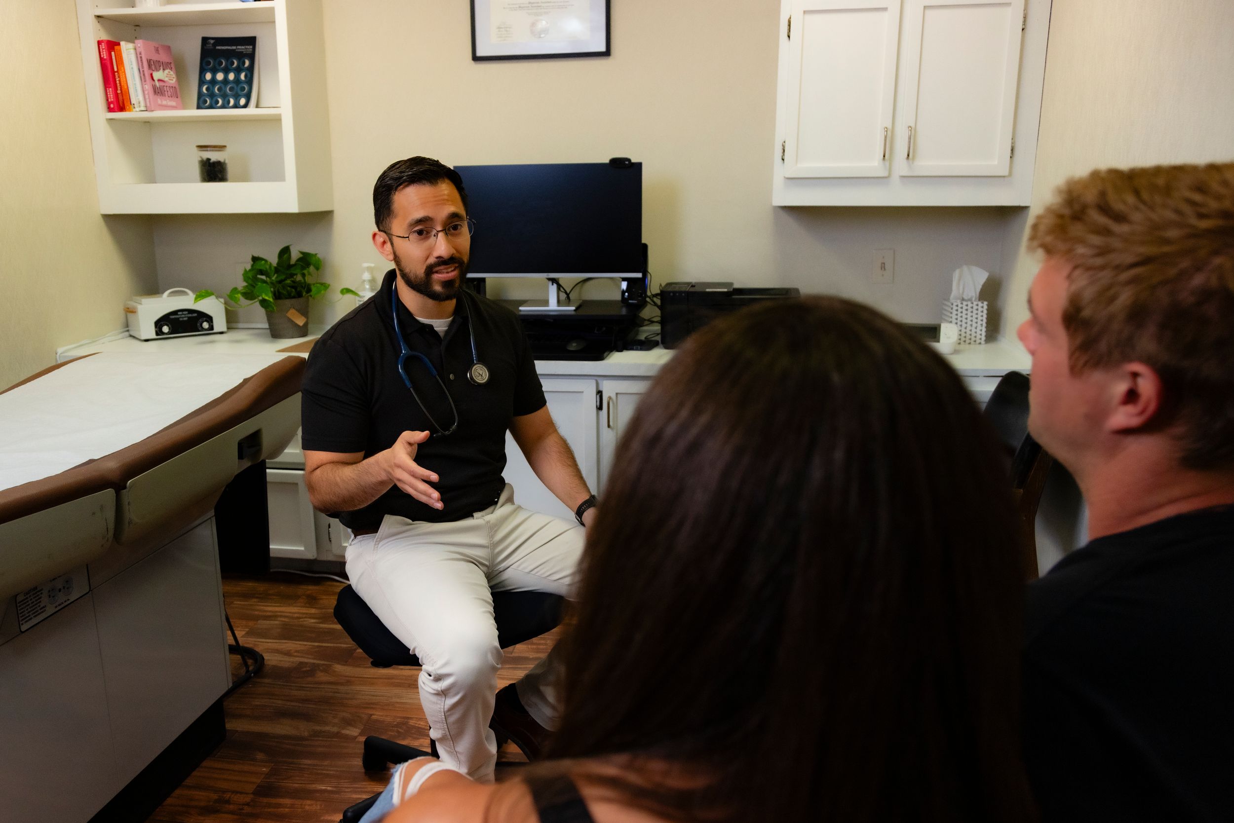 Healthcare provider sits at desk speaking with patients during medical consultation in examination room.