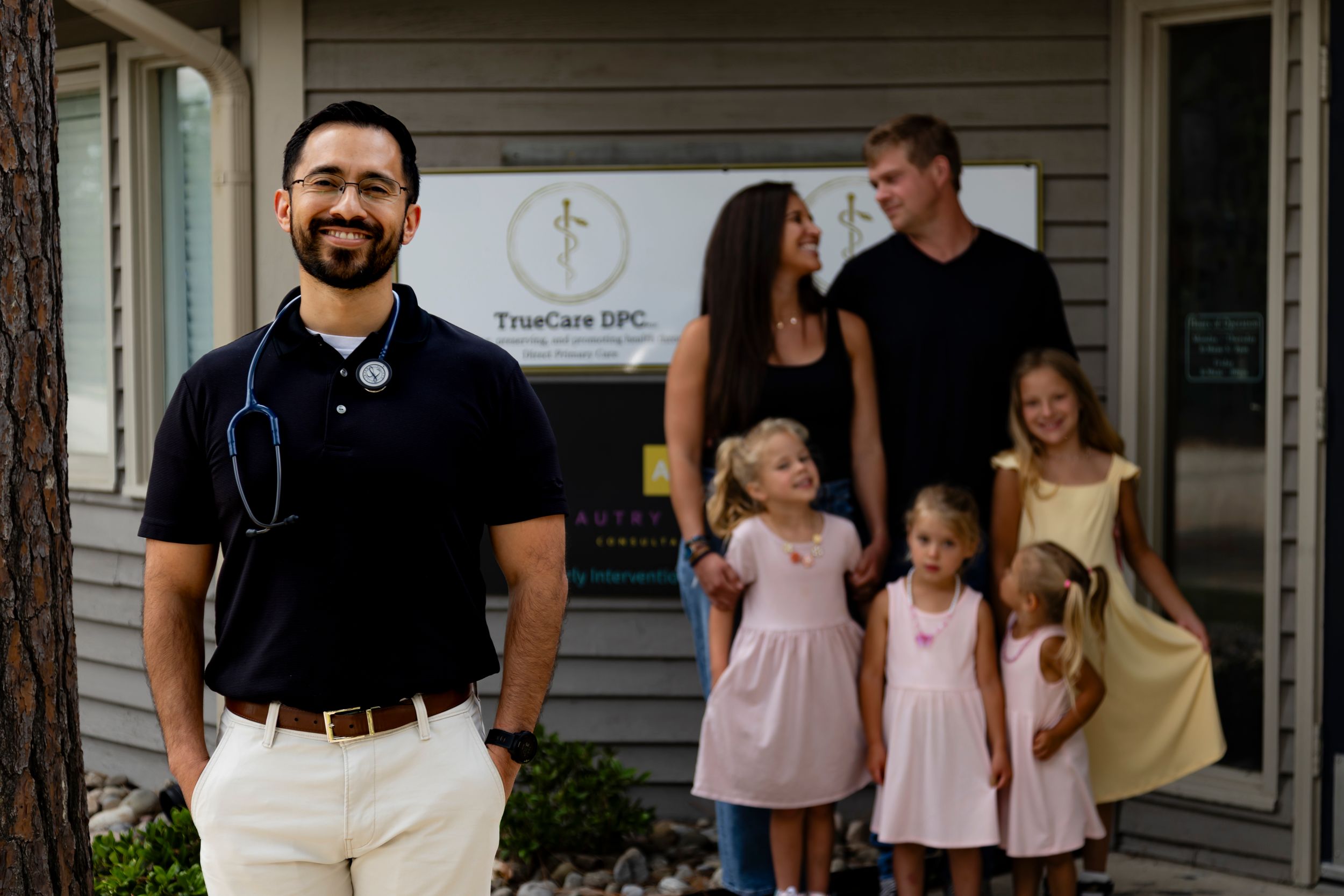 People stand outside the entrance of a clinic or medical office building with a sign visible on the exterior wall.