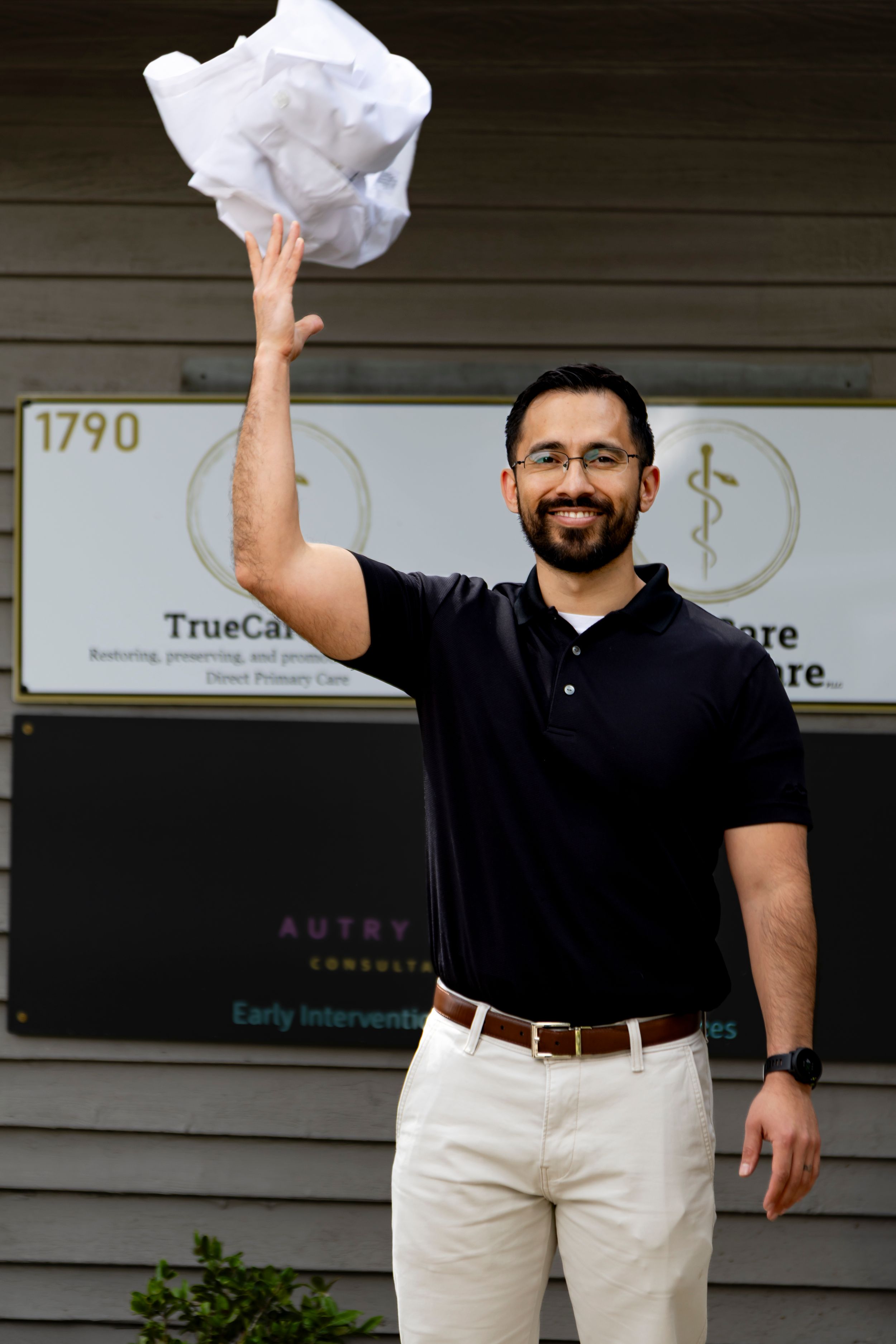 Man wearing black polo shirt and khaki pants waves while standing in front of business office.