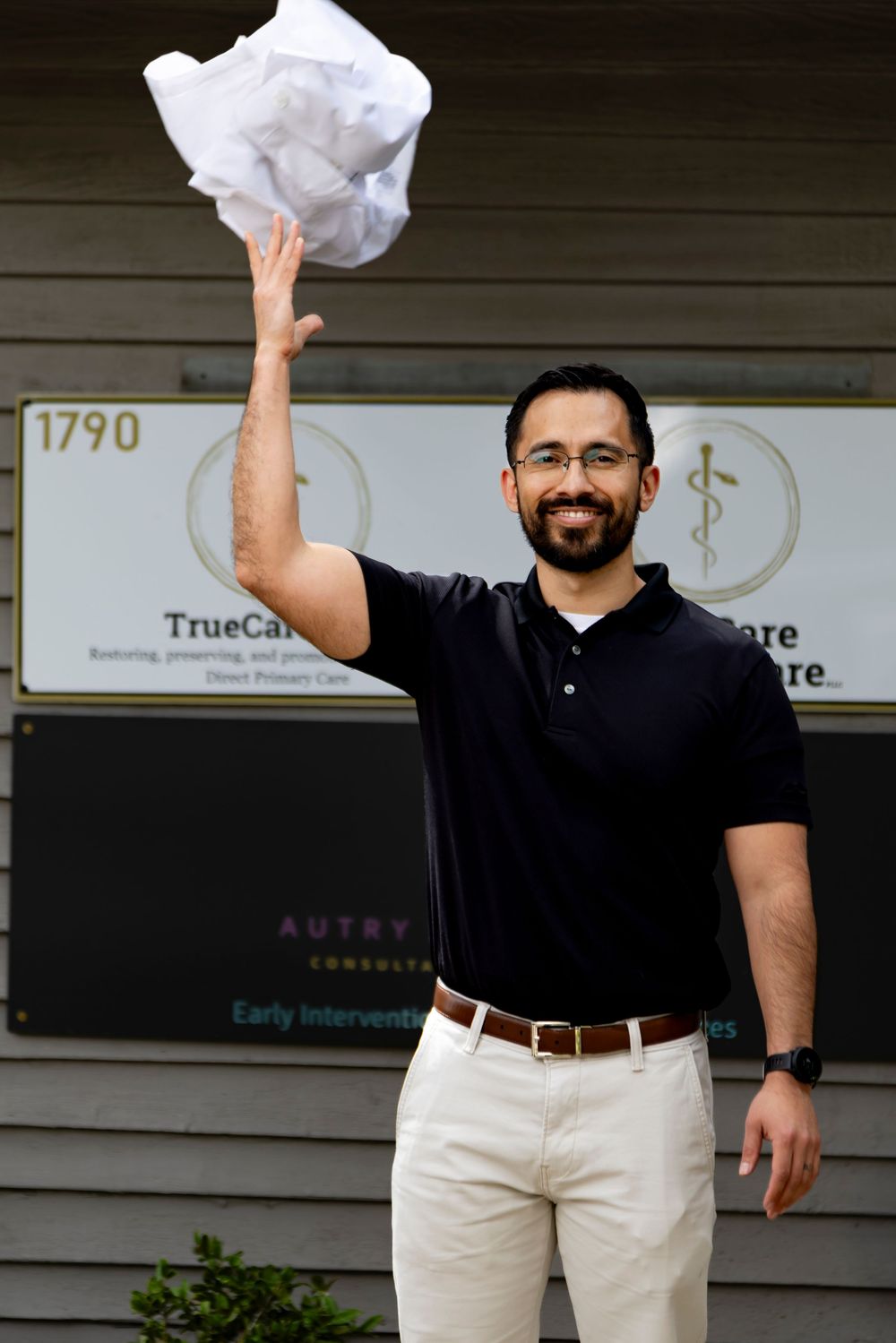 Man wearing black polo shirt and khaki pants waves while standing in front of business office.