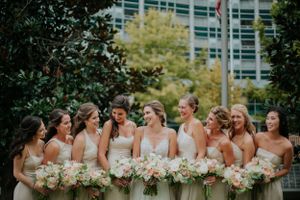 Bridesmaids in light colored dresses stand together holding pink and white bouquets outdoors.