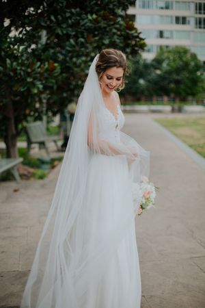 Bride in flowing white gown and long veil holds bouquet, smiling outdoors near trees on her wedding day.