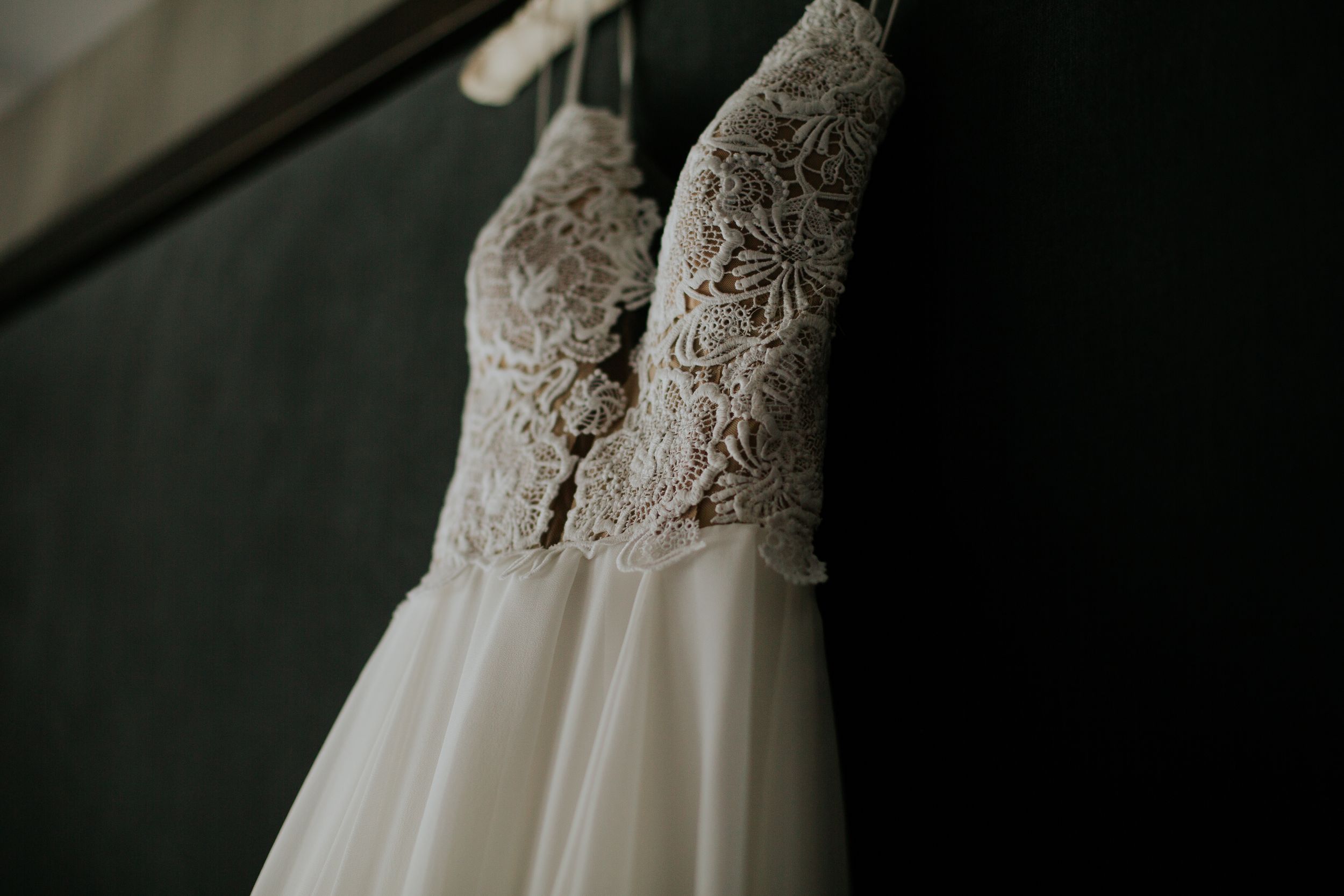 Close-up detail shot of an elegant white lace wedding dress bodice hanging against a dark background.