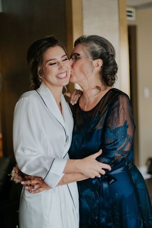 A bride in a white robe smiles as an older woman in a blue dress kisses her cheek during wedding preparations.