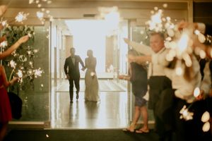 Wedding guests celebrate as silhouettes enter through bright doorway with sparkling lights in foreground.
