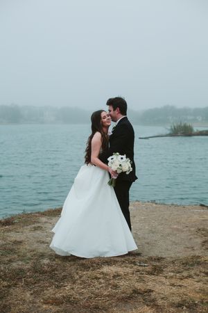 Wedding couple embraces near misty lake holding white bouquet in romantic coastal setting.