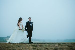A couple in wedding attire stands on cliff edge overlooking foggy water landscape.