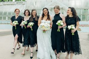 Bride in white gown with five bridesmaids in black dresses holding white bouquets, walking outside a greenhouse.