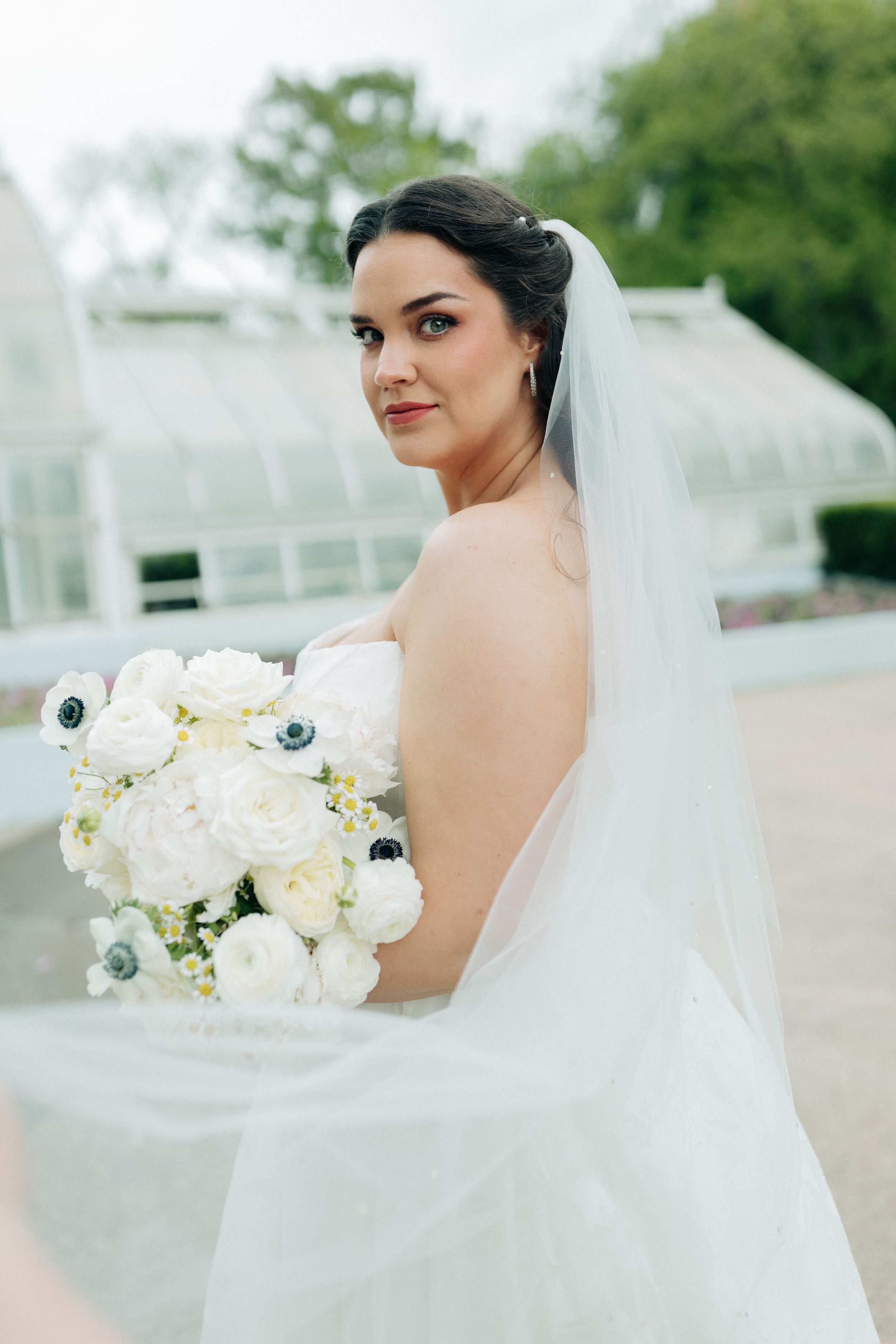 Bride in backless white gown holds bouquet of white ranunculus and anemones, veil flowing, posing outdoors near greenhouse.