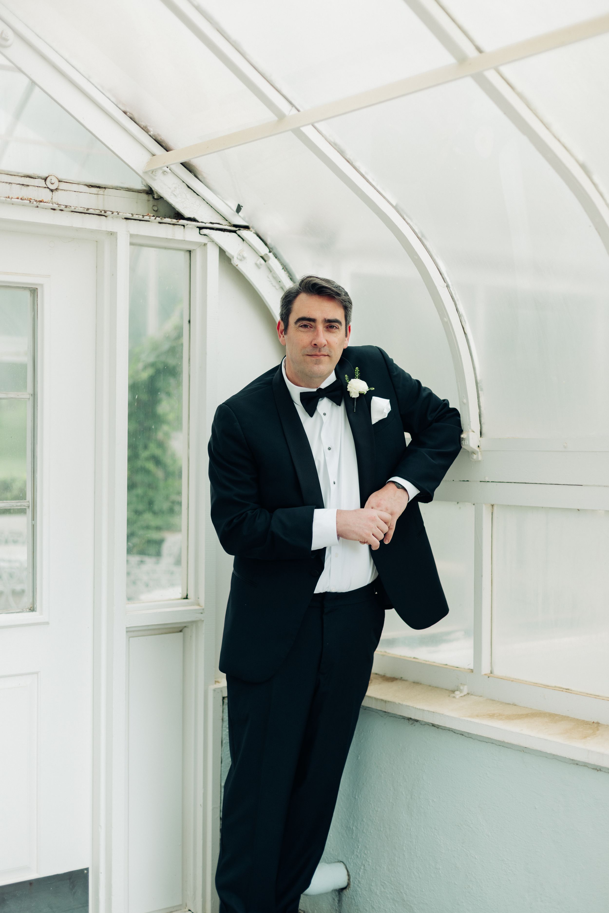Groom in black tuxedo with bow tie and boutonniere posing in elegant greenhouse venue on wedding day.