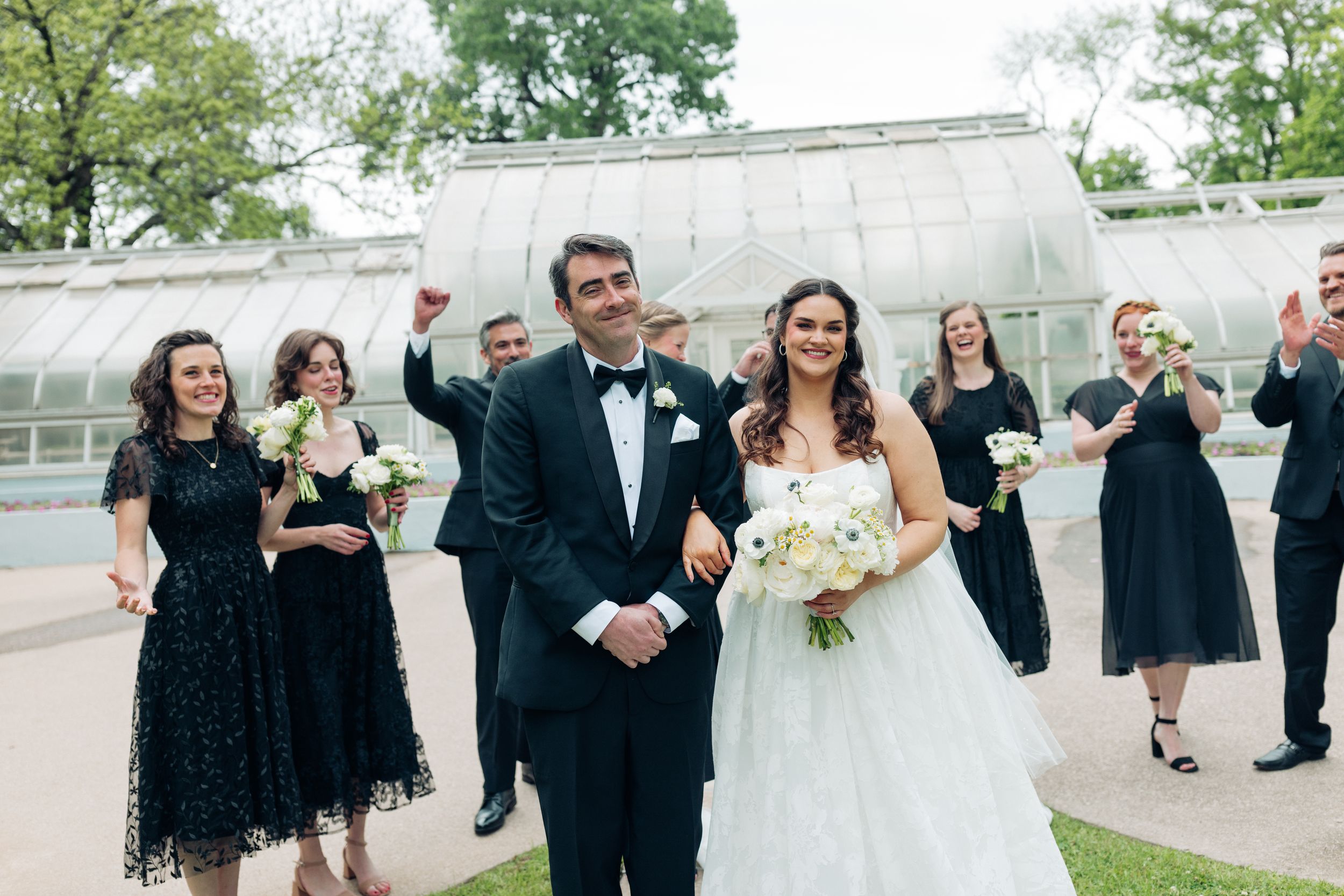Bride and groom walk joyfully as wedding party in black attire celebrates behind them near a greenhouse.