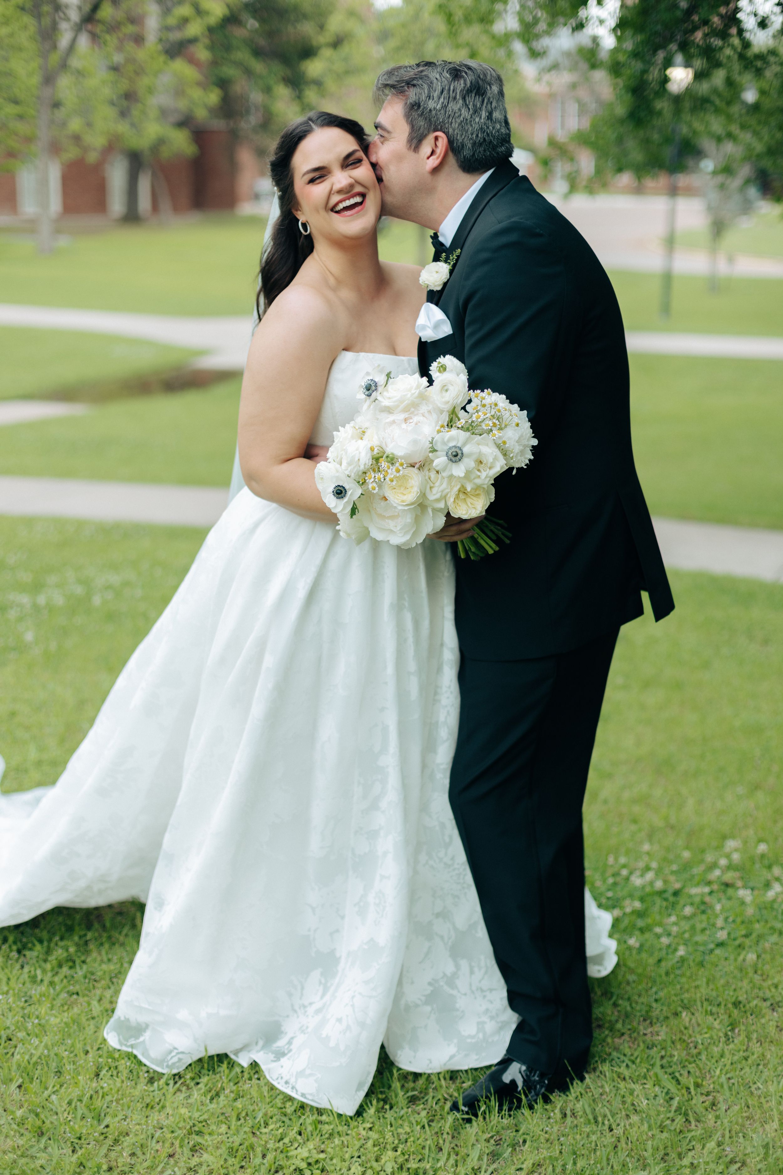 Bride in strapless white gown holding bouquet smiles as groom in black suit kisses her cheek outdoors.