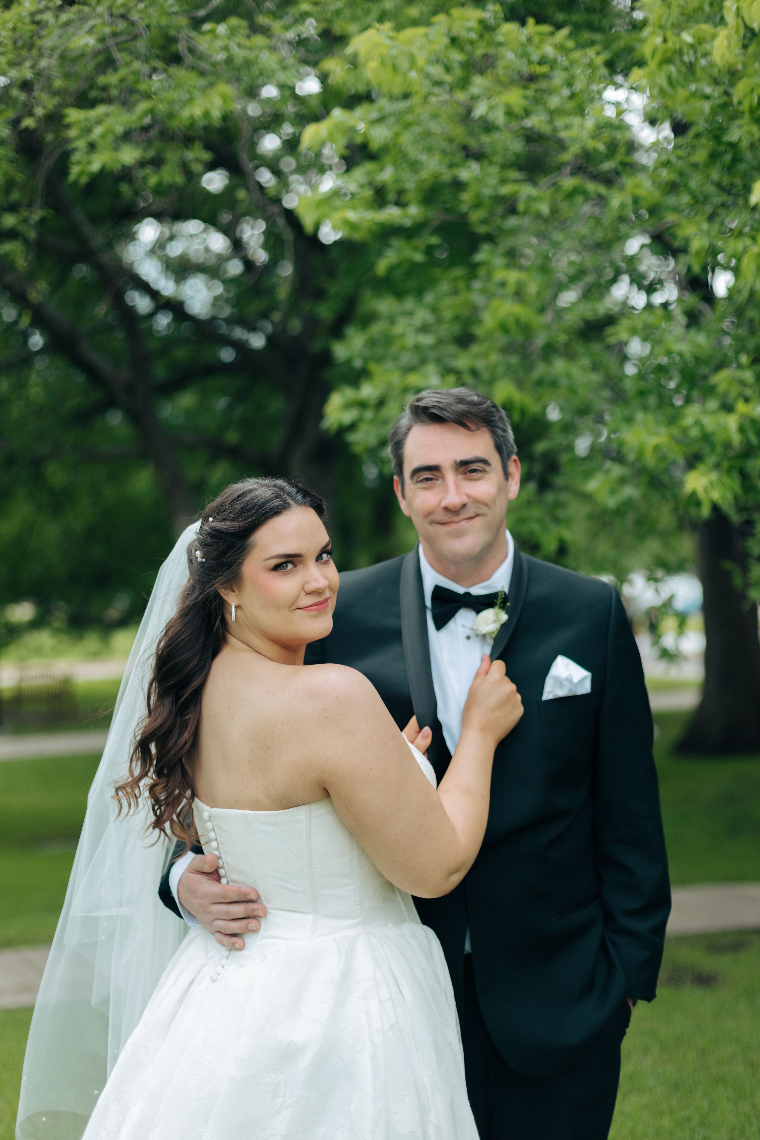 Bride in strapless white gown and groom in black tuxedo pose together outdoors with lush green trees behind them.