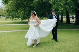 Bride in white gown twirling while groom in tuxedo assists with dress train on green lawn.