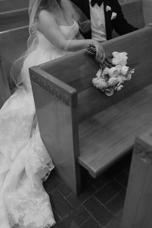 Black and white photo of a bride and groom sitting in a church pew with a white floral bouquet resting beside them.