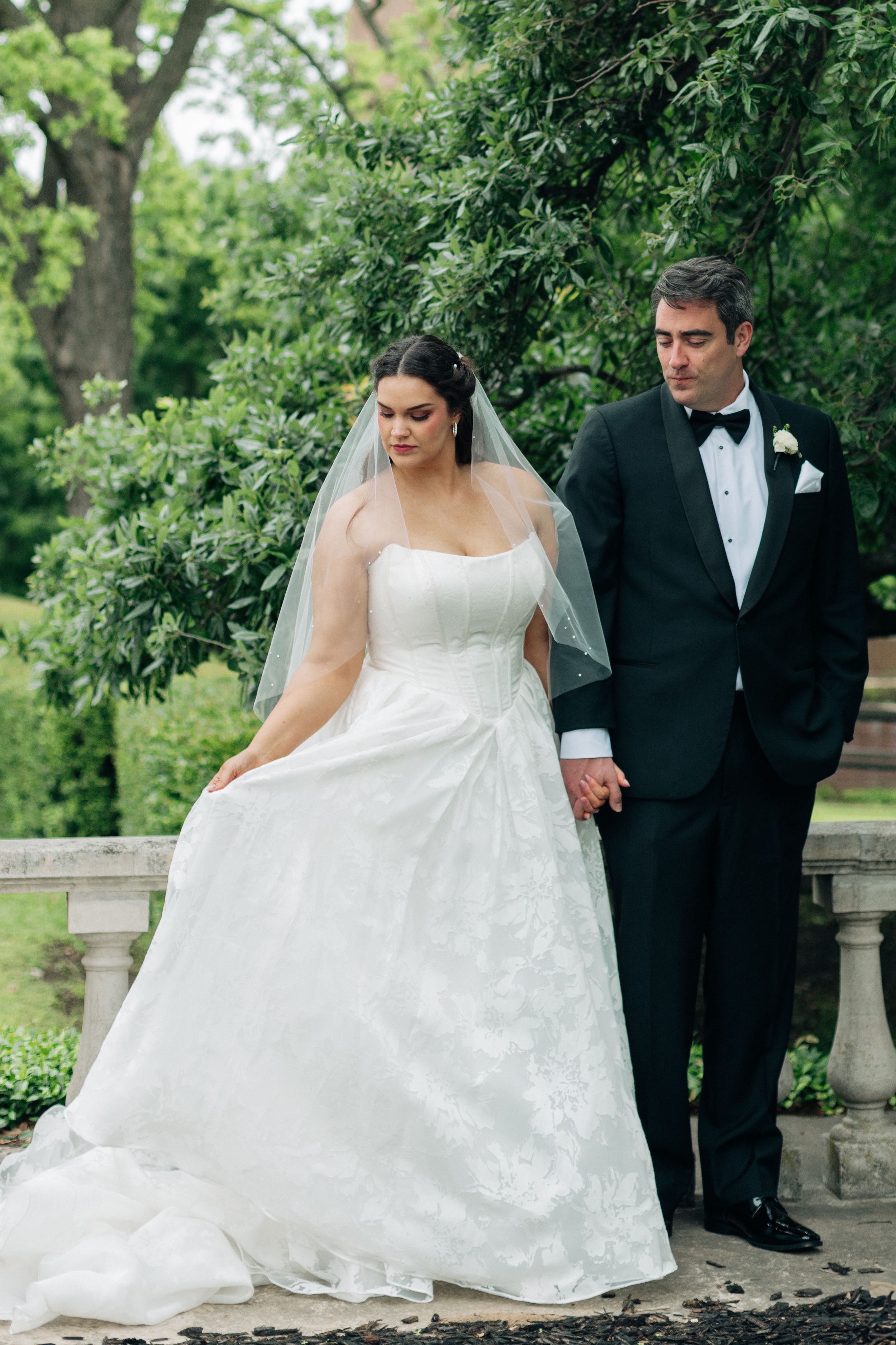 Bride in white lace ball gown and veil holds hands with groom in black tuxedo in lush garden setting.