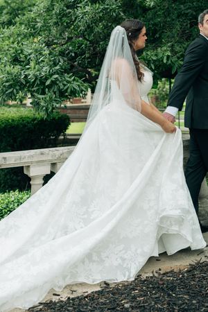 Bride in elegant lace wedding gown with long veil walks with groom in garden setting.