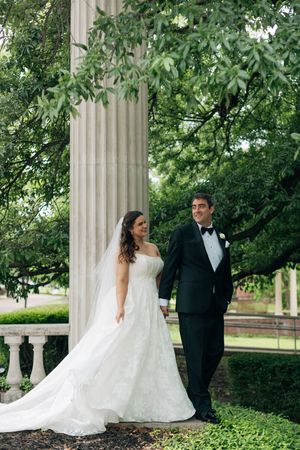 Bride in white lace gown and veil stands with groom in black tuxedo beside stone column surrounded by lush greenery.