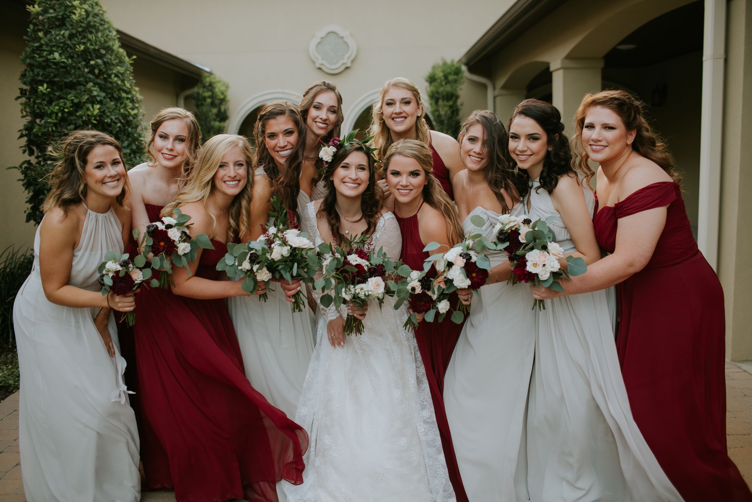 Bridal party in burgundy and white dresses pose together with bouquets.
