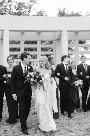Black and white photo of a wedding party walking together under a white pergola structure.