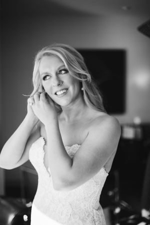 A bride wearing a white strapless lace gown touches her earring while smiling in a black and white portrait.