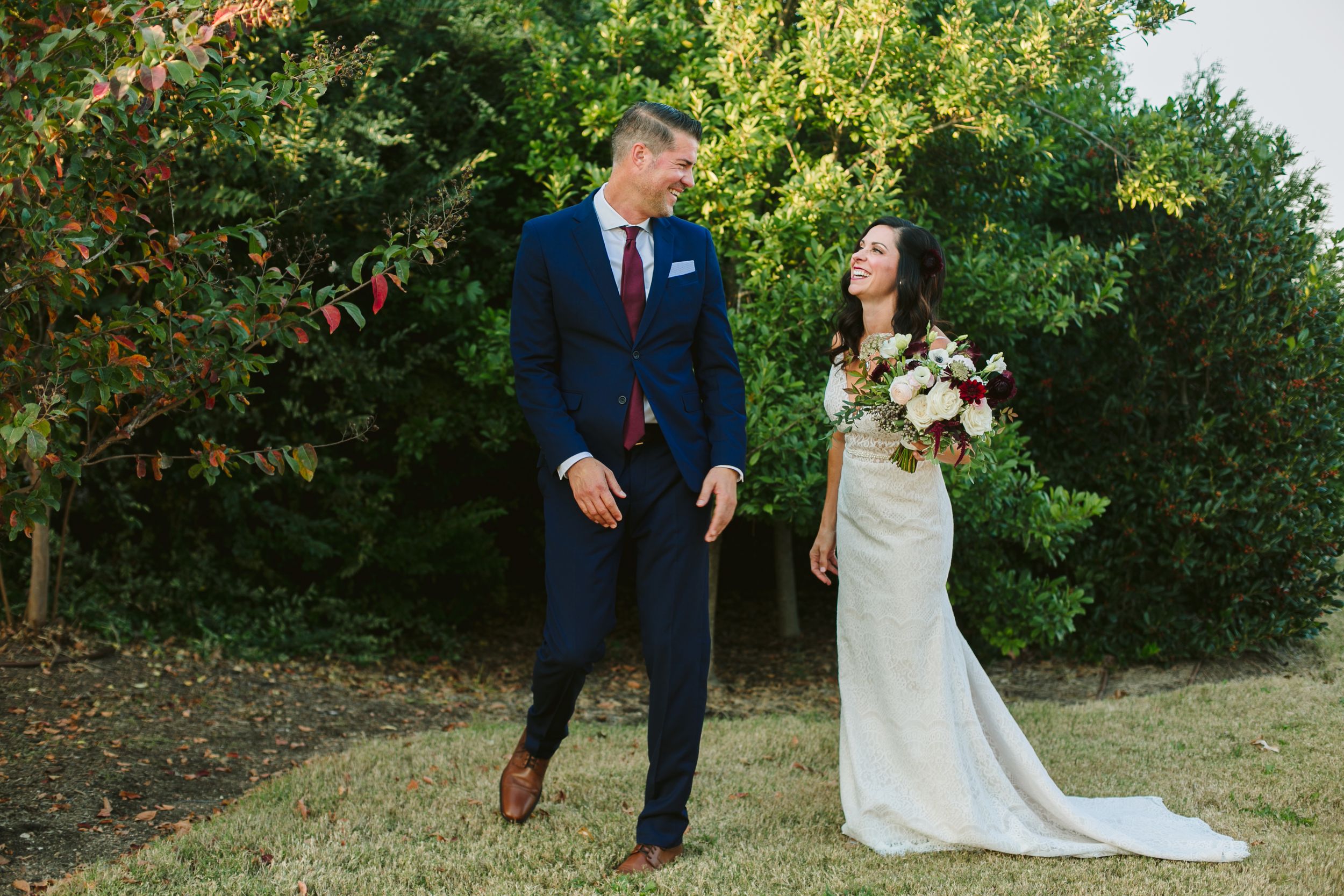 Couple shares a candid moment outdoors with bride holding a lush bouquet against green foliage.
