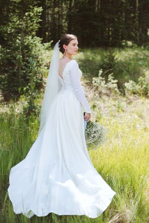 Bride in flowing long-sleeve gown stands in sunlit meadow with forest backdrop.