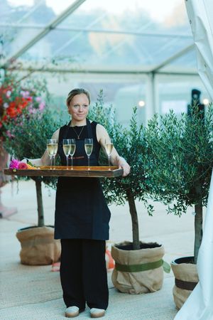A waitress in black attire carries a wooden tray with champagne glasses at an elegant outdoor event.
