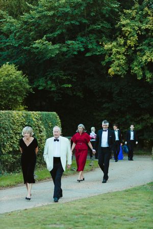 Elegantly dressed couples in formal attire walk along a garden path surrounded by lush green trees.
