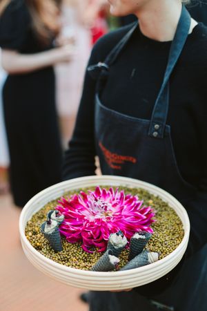 Chef holding a round tray with pink dahlia flower, small cones, and seeds at a catering event.