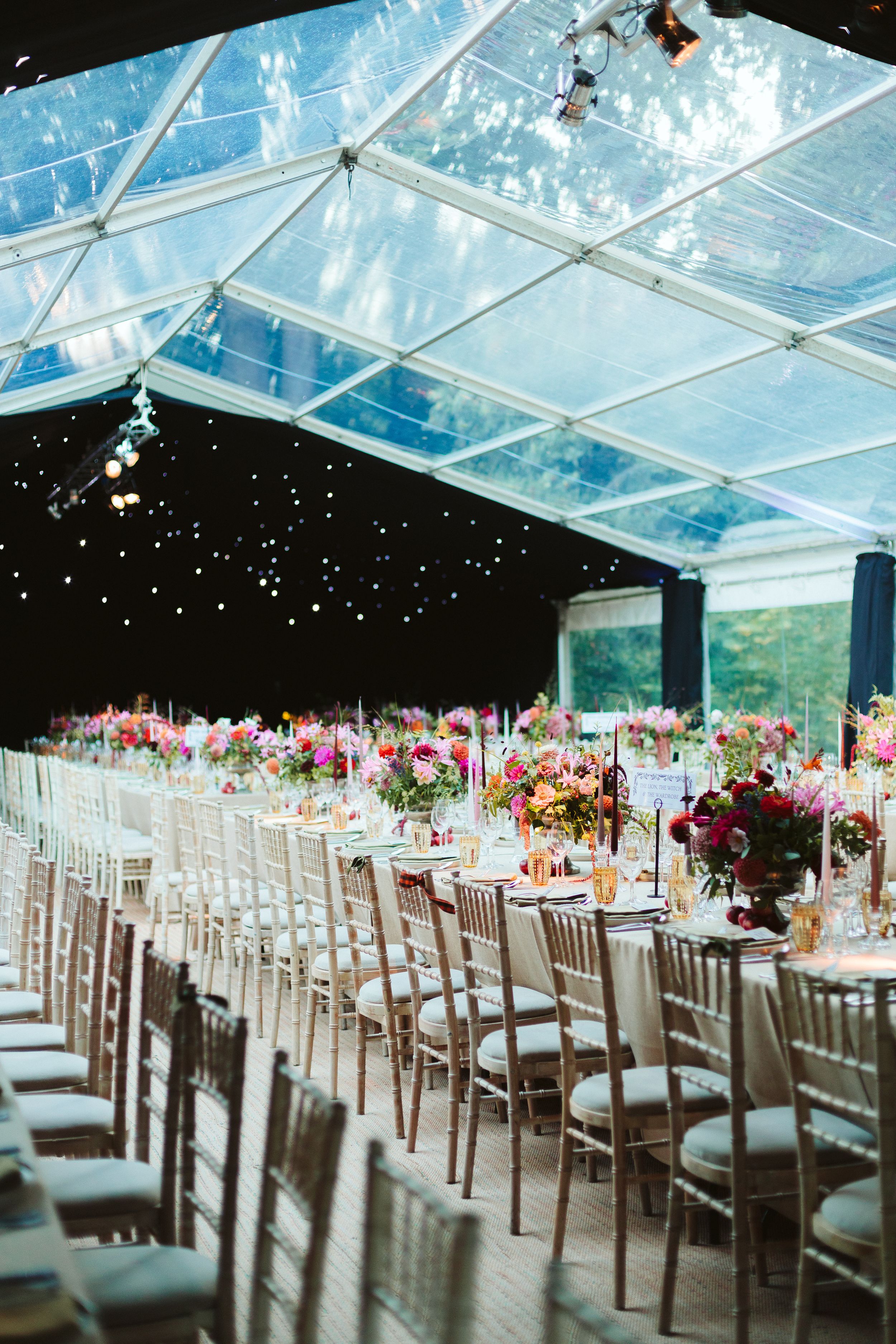 Elegant wedding reception in a clear marquee tent with long tables, chiavari chairs, and vibrant floral centerpieces.