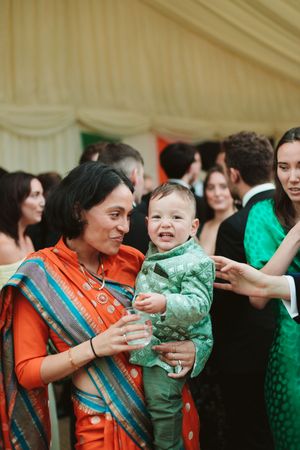 Woman in orange sari holds smiling baby in green outfit at elegant indoor celebration.