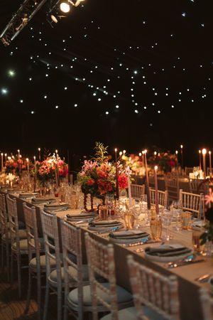 Elegant wedding reception with twinkling lights, floral centerpieces, candles, and silver chiavari chairs at long dining tables.