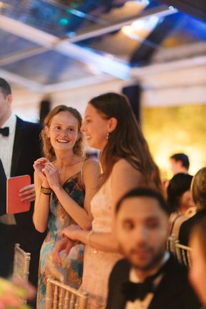 Two elegantly dressed women smiling and clapping at a formal evening gala event under a tented venue.