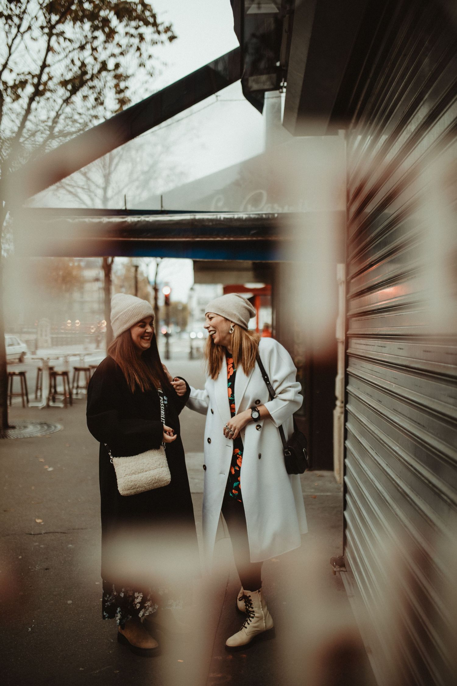 Two friends in winter coats and beanies chat on a city street beside a closed storefront shutter.