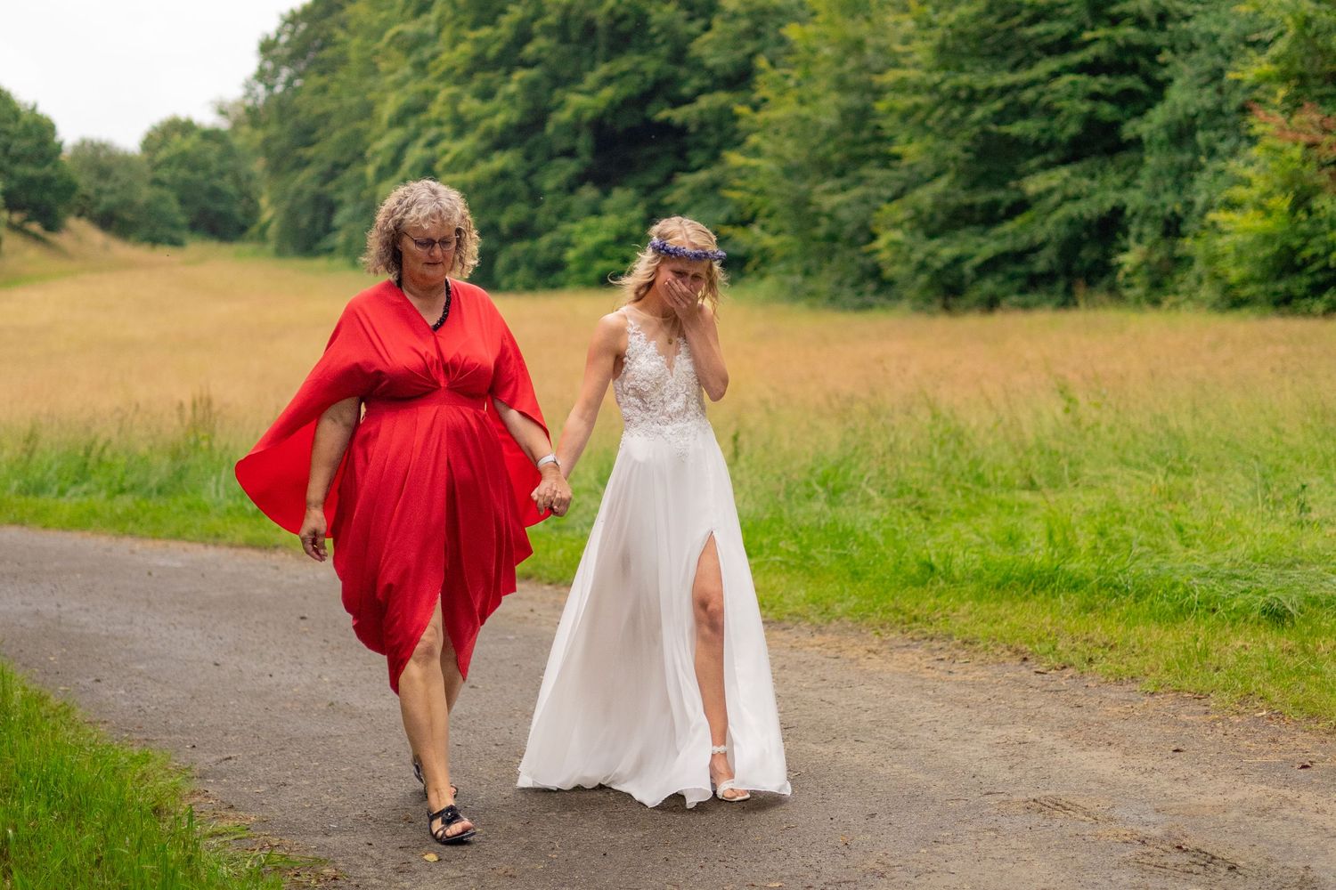 Two women walking together on a countryside path, one wearing red and one in a white wedding gown.