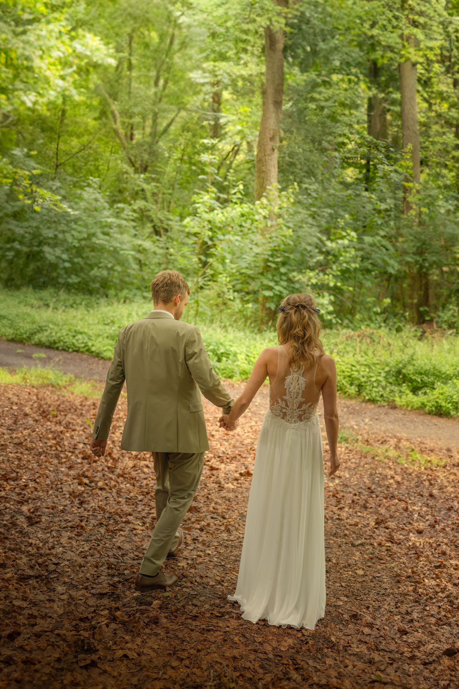 Newlyweds walking hand in hand down a leaf-covered forest path in ethereal lighting.