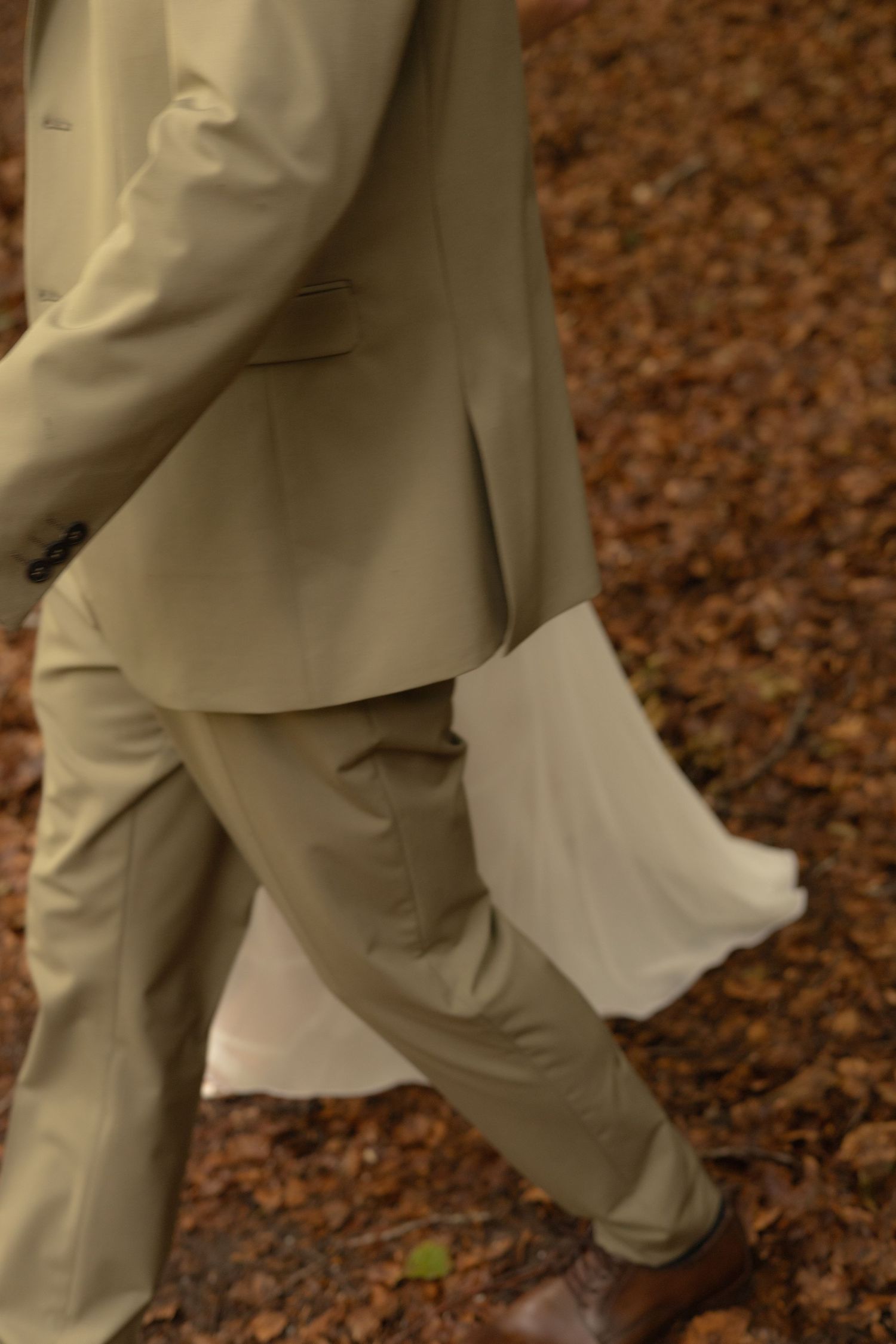 Close up of khaki suit pants and dress shoes walking on ground covered in brown leaves.