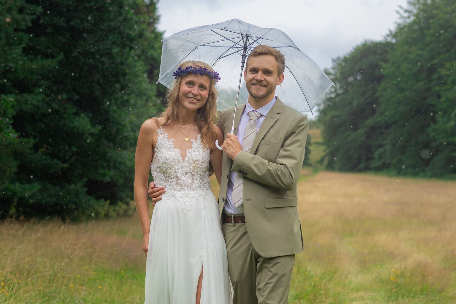 Wedding couple standing together under clear umbrella in a rainy meadow.