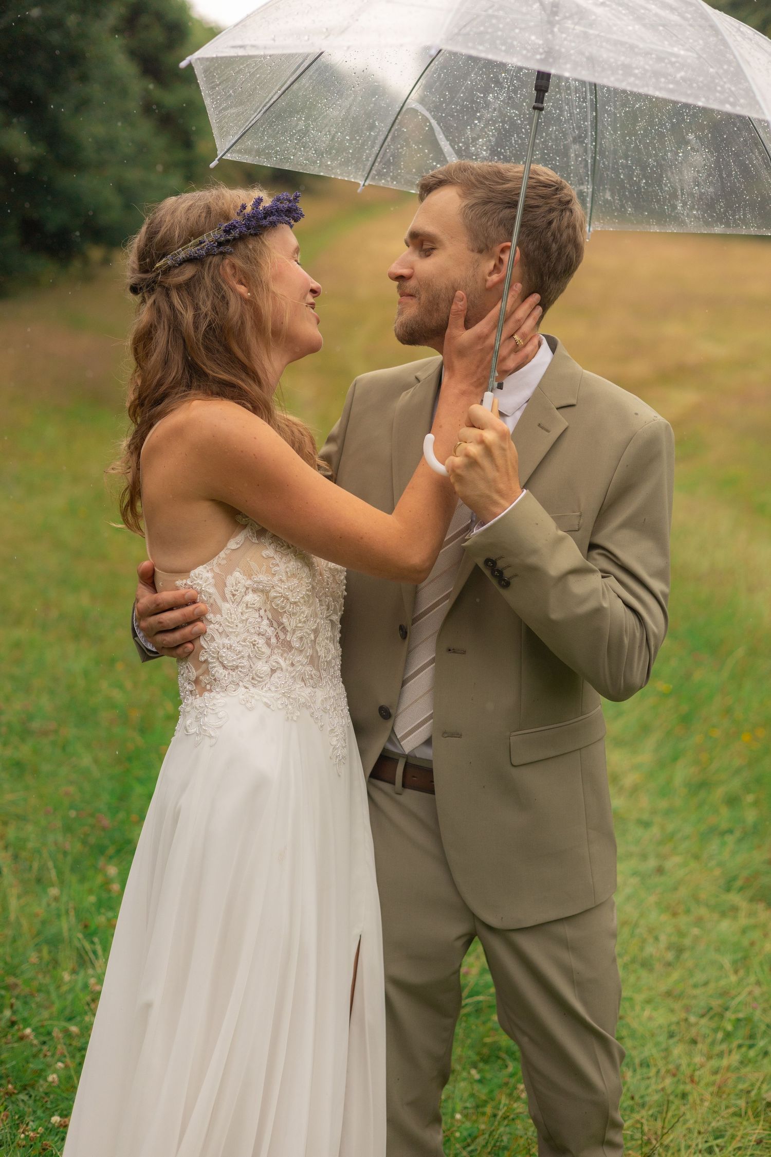 Romantic wedding couple sharing intimate moment under clear umbrella in meadow.