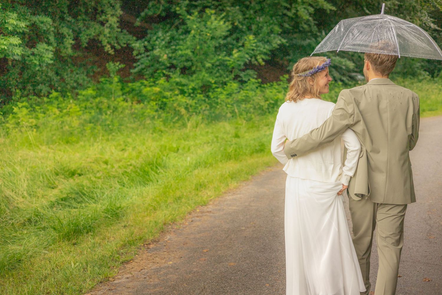 Newlyweds walking away on path under clear umbrella surrounded by greenery.